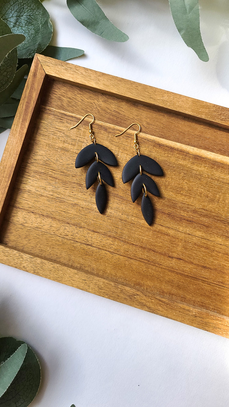 Solid black earrings in a three piece petal design. The earrings lay against a wooden tray and eucalyptus leaves frame the image.